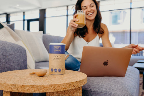 A woman in front of a laptop, holding an iced latte, next to the product in a cylinder carton packaging..
